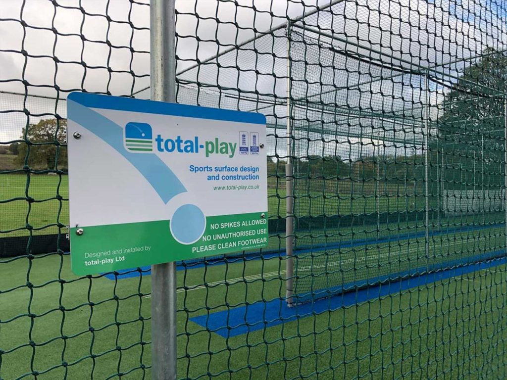 A board on display on a fence with cricket netting outside a cricket turf.